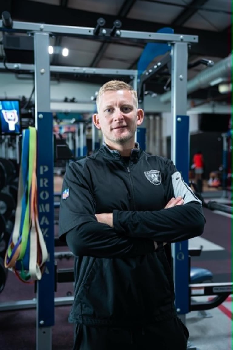 Man in black Raiders jacket posing with arms crossed inside a modern gym facility with exercise equipment